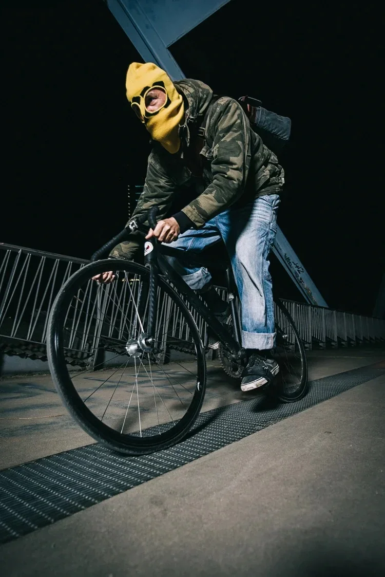 Cyclist in a yellow balaclava riding across a bridge at night captured with high-contrast flash.